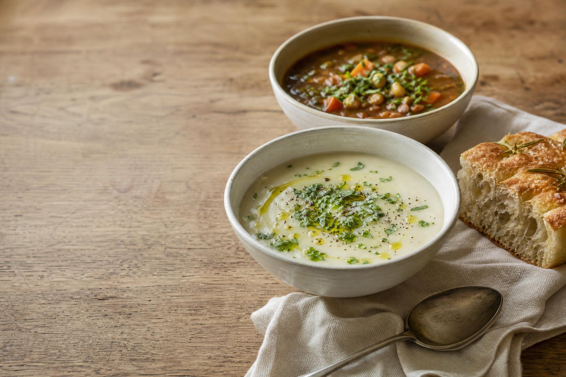 Två soppskålar på ett rustikt träbord, en ljus krämig vegetarisk soppa och en fylligare soppa, med en bit nybakad focaccia